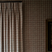 A close-up of a pleated curtain panel hanging on a dark rod against a matching patterned wallpaper with a subtle grid design.